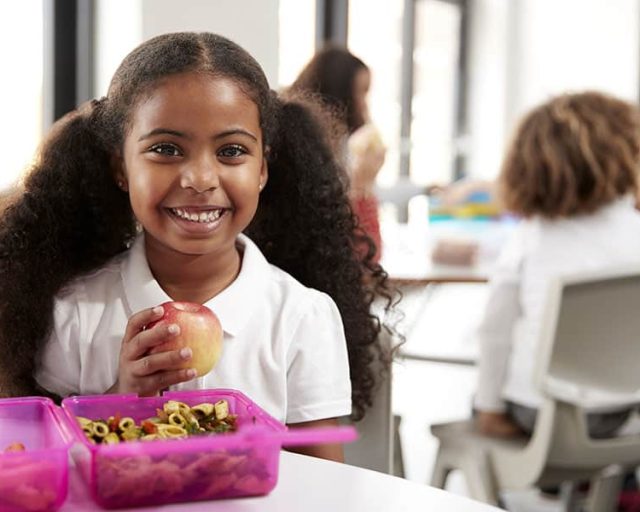 Young black schoolgirl sitting at a table smiling and holding an apple in a kindergarten classroom during her lunch break, close up