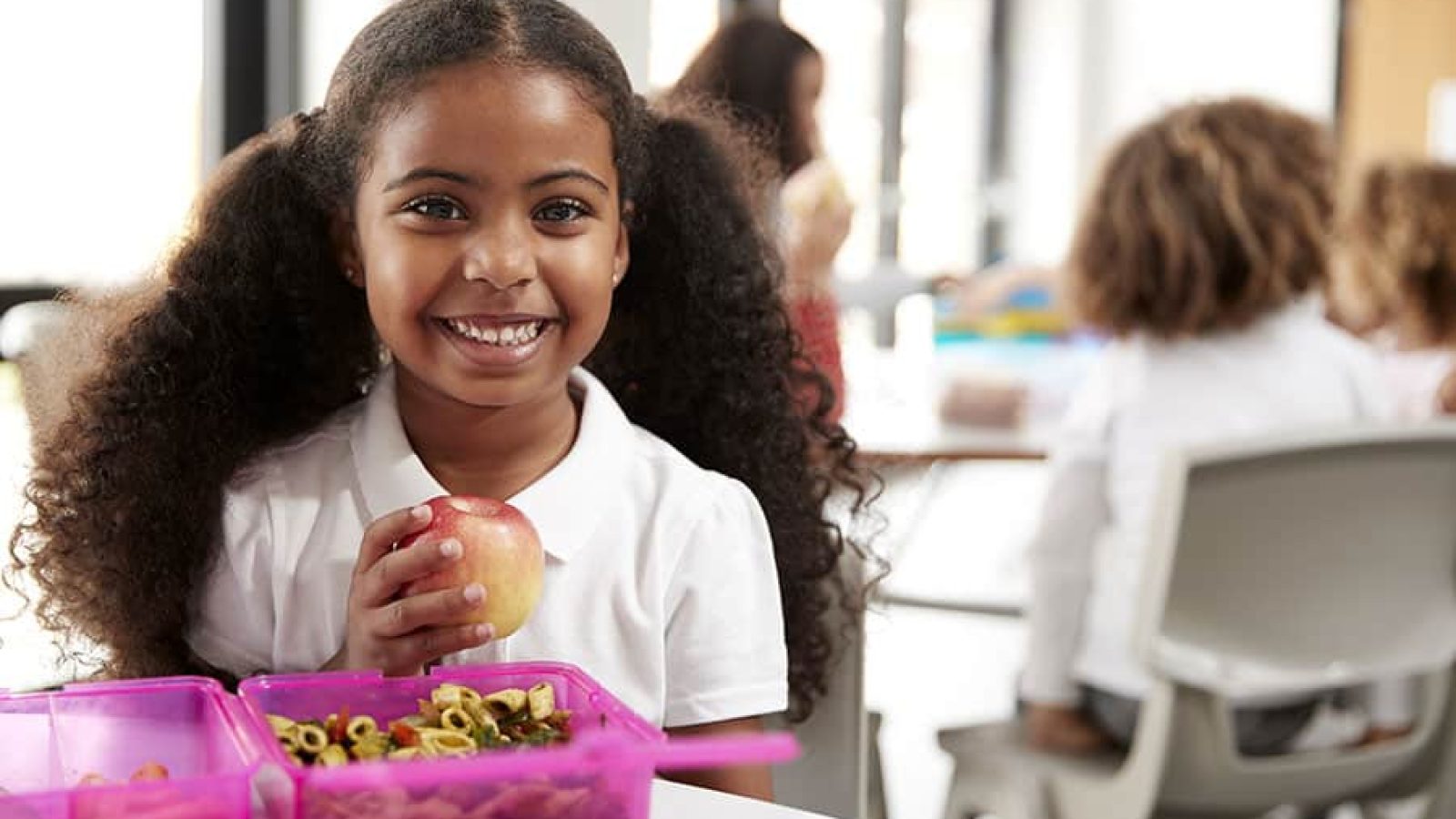Young black schoolgirl sitting at a table smiling and holding an apple in a kindergarten classroom during her lunch break, close up